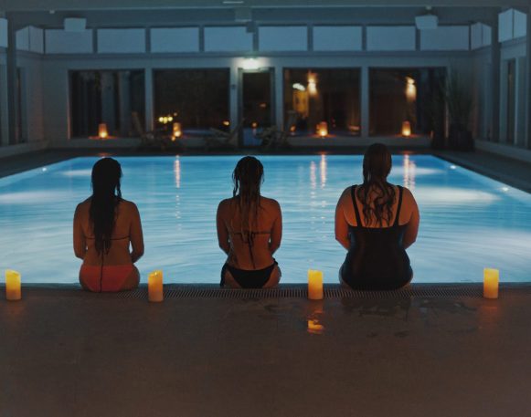 tre kvinner sitter med ryggen mot kamera på en bassengkant, med levende lys mellom seg