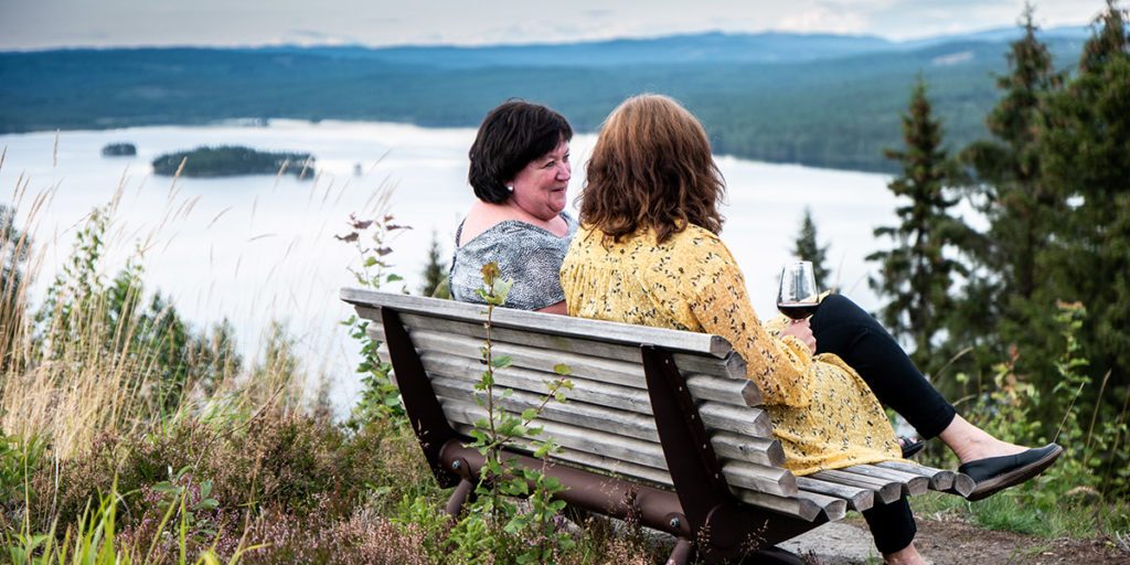 to kvinner sitter på en benk og snakker sammen med et vinglass i hånden, og helvt vendt fra kamera, med en sjø og skog i bakgrunnen