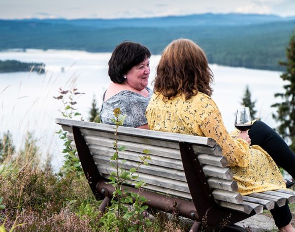 to kvinner sitter på en benk og snakker sammen med et vinglass i hånden, og helvt vendt fra kamera, med en sjø og skog i bakgrunnen