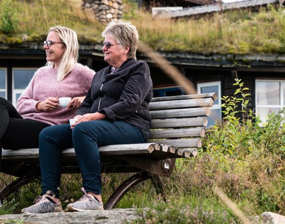 to kvinner sitter på en benk og snakker sammen med et kaffekopp i hånden, og halvt vendt fra kamera, med finnskogtoppen i bakgrunnen
