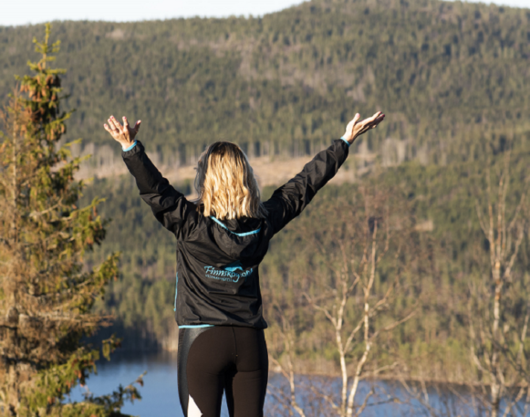 En kvinne står med armene hevet og ryggen mot kamera, og ser utover Finnskogen