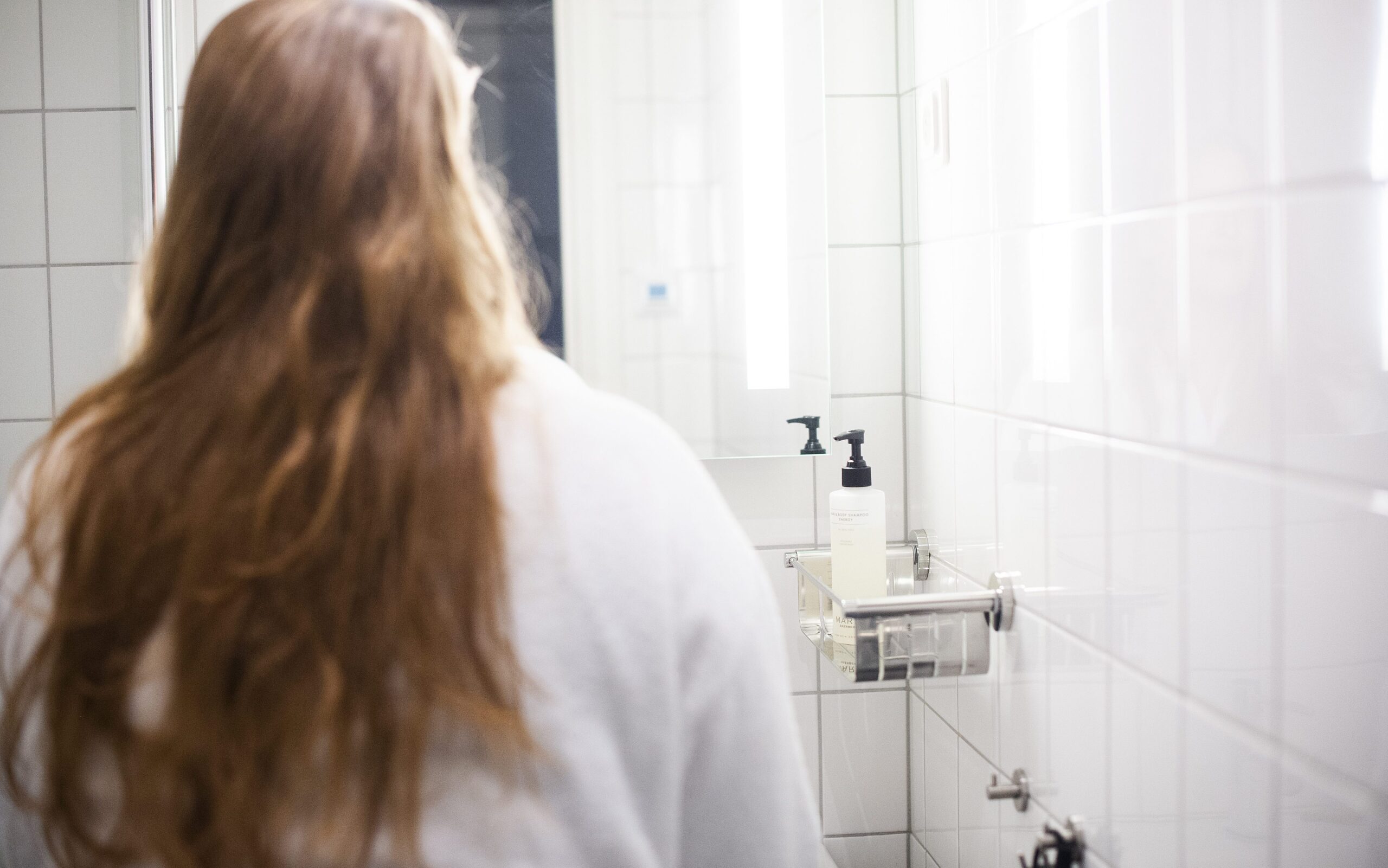 En kvinne med langt lyst hår og badekåpe står på et bad med ryggen til kamera