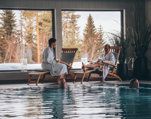 Bildet viser to personer ikledd morgenkåper i hver sin stol på bassengkanten i bakgrunnen. I forgrunnen svømmer to personer i bassenget på Finnskogtoppen Spa og Velværehotell.